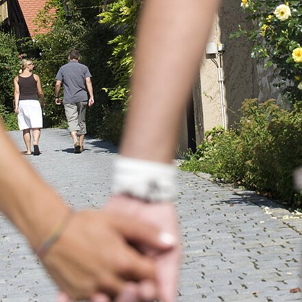 Paar hält Händchen im Vordergrund, zwei Personen gehen auf gepflastertem Weg an Fachwerkhäusern entlang.