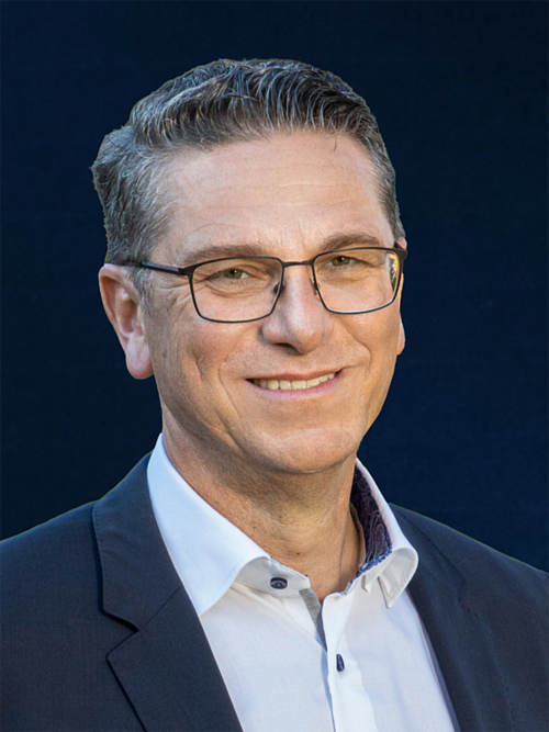 Porträt eines lächelnden Mannes mit Brille, weißem Hemd und dunklem Sakko vor dunklem Hintergrund.