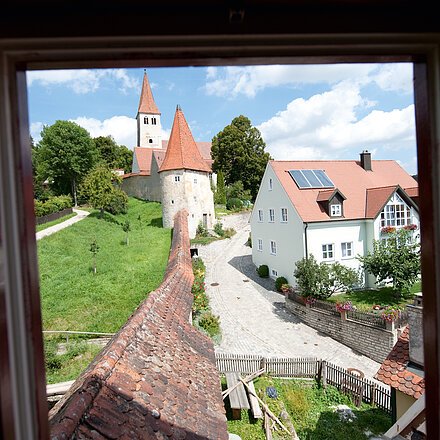 Blick aus Fenster auf gepflasterte Straße, Häuser mit roten Dächern und eine Kirche mit Turm im Hintergrund.
