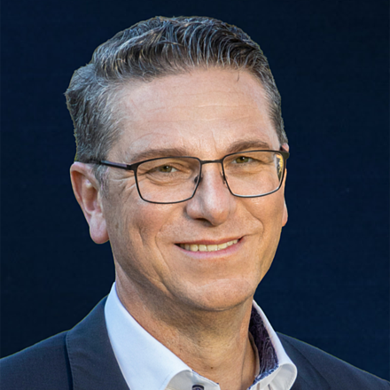 Porträt eines lächelnden Mannes mit Brille, weißem Hemd und dunklem Sakko vor dunklem Hintergrund.