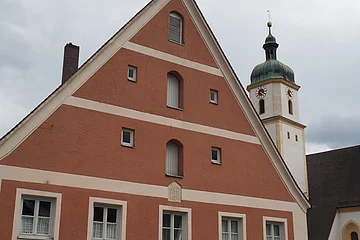 Dreistöckiges rotes Haus mit weißen Fensterrahmen und Kirchturm mit grünem Dach im Hintergrund bei bewölktem Himmel.
