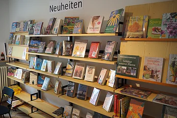 Regal mit verschiedenen Büchern und Schild „Neuheiten“ in einem Raum mit Stuhl und Heizkörper.