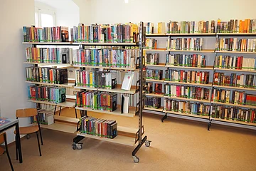 Regal mit vielen Büchern in einem hellen Raum mit Stuhl und Tisch links.