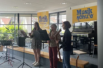 Drei Personen spielen Blockflöte auf einer Bühne vor Fenstern mit zwei gelben Musikschule-Bannern.