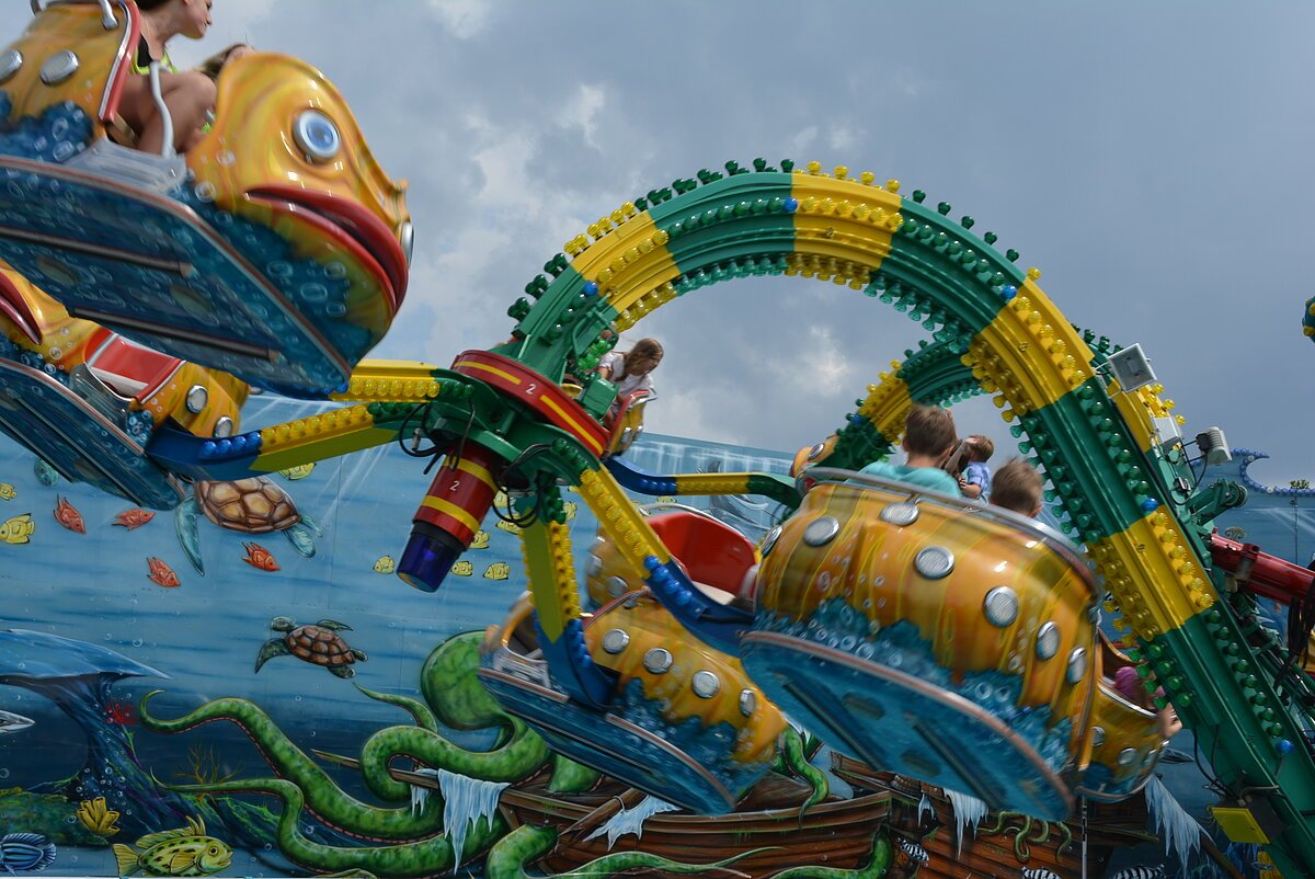 Fahrgeschäft mit gelb-blauen Fischwagen und grüner-gelber Schienenstruktur vor bemalter Wand mit Meeresmotiven.