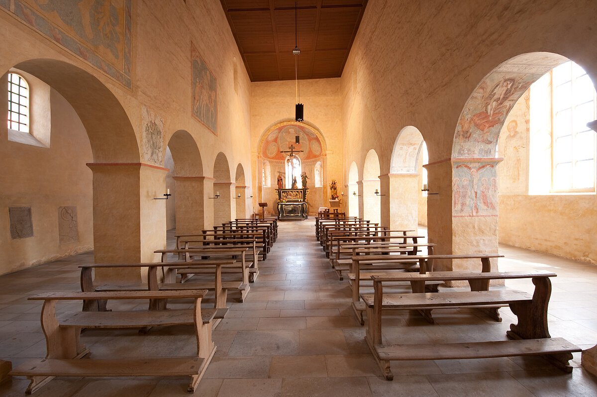 Innenraum einer Kirche mit Holzbänken, Säulen und Altar im Hintergrund, helle Wände mit Fresken und Fenstern