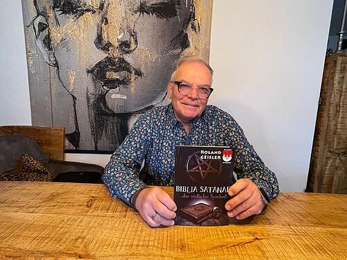 Mann mit Brille sitzt an Holztisch und hält Buch „Biblia Satanae“ von Roland Geisler hoch.