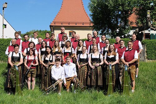 Gruppenfoto einer Blaskapelle mit 31 Personen in Trachten vor Haus mit rotem Dach bei Sonnenschein auf Wiese.