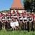 Gruppenfoto einer Blaskapelle mit 31 Personen in Trachten vor Haus mit rotem Dach bei Sonnenschein auf Wiese.