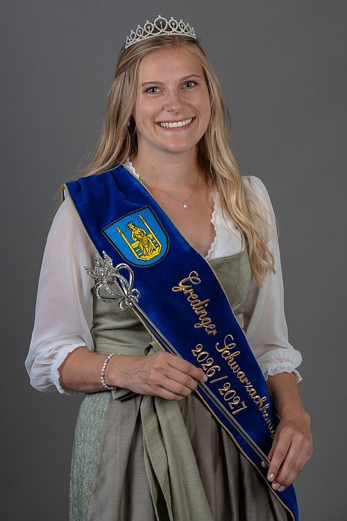 Gredinger Schwarzachkönigin 2026/2027 Gredinger Schwarzachkönigin 2026/2027 mit Diadem, blauem Schärpenband und Zepter vor grauem Hintergrund im Dirndl.