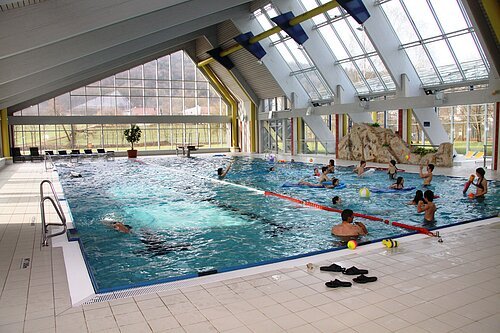Innenaufnahme eines Schwimmbads mit Schwimmern und spielenden Personen im Wasser, große Fensterfront sichtbar.
